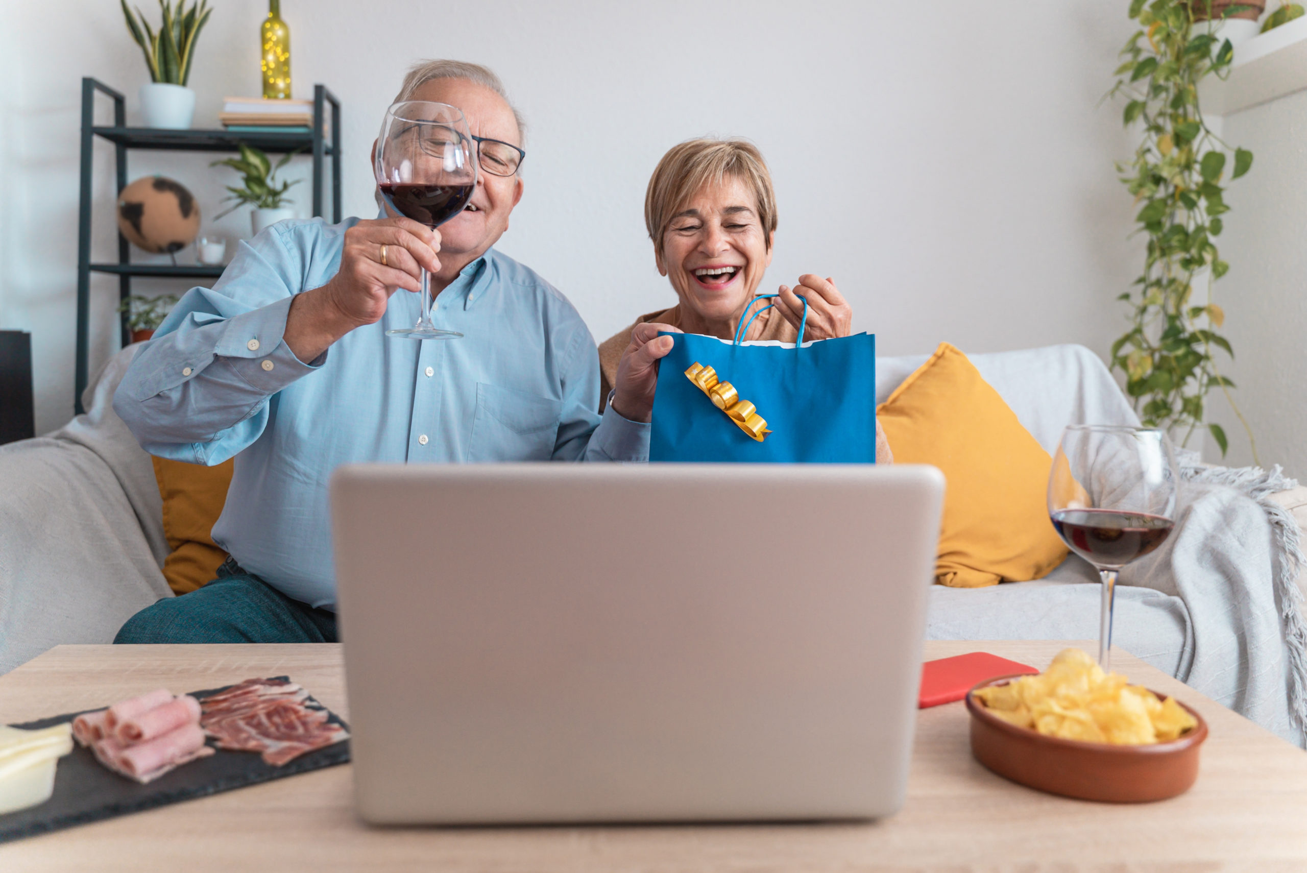 Happy senior couple celebrating with a toast with wine on video call to celebrate christmas or birthday during coronavirus outbreak - Tecnology and family party concept - Focus on gift