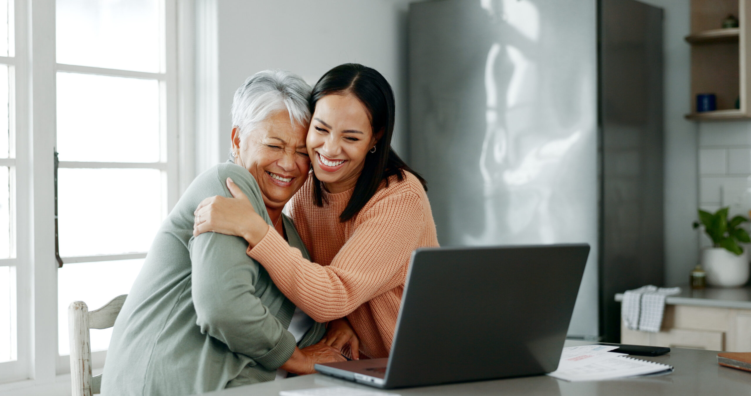A senior woman with short gray hair and a younger woman with long dark hair share a warm embrace while smiling. They are sitting in a bright kitchen with a laptop open on the counter, surrounded by papers. Natural light streams in through large windows, creating a cozy and happy atmosphere.