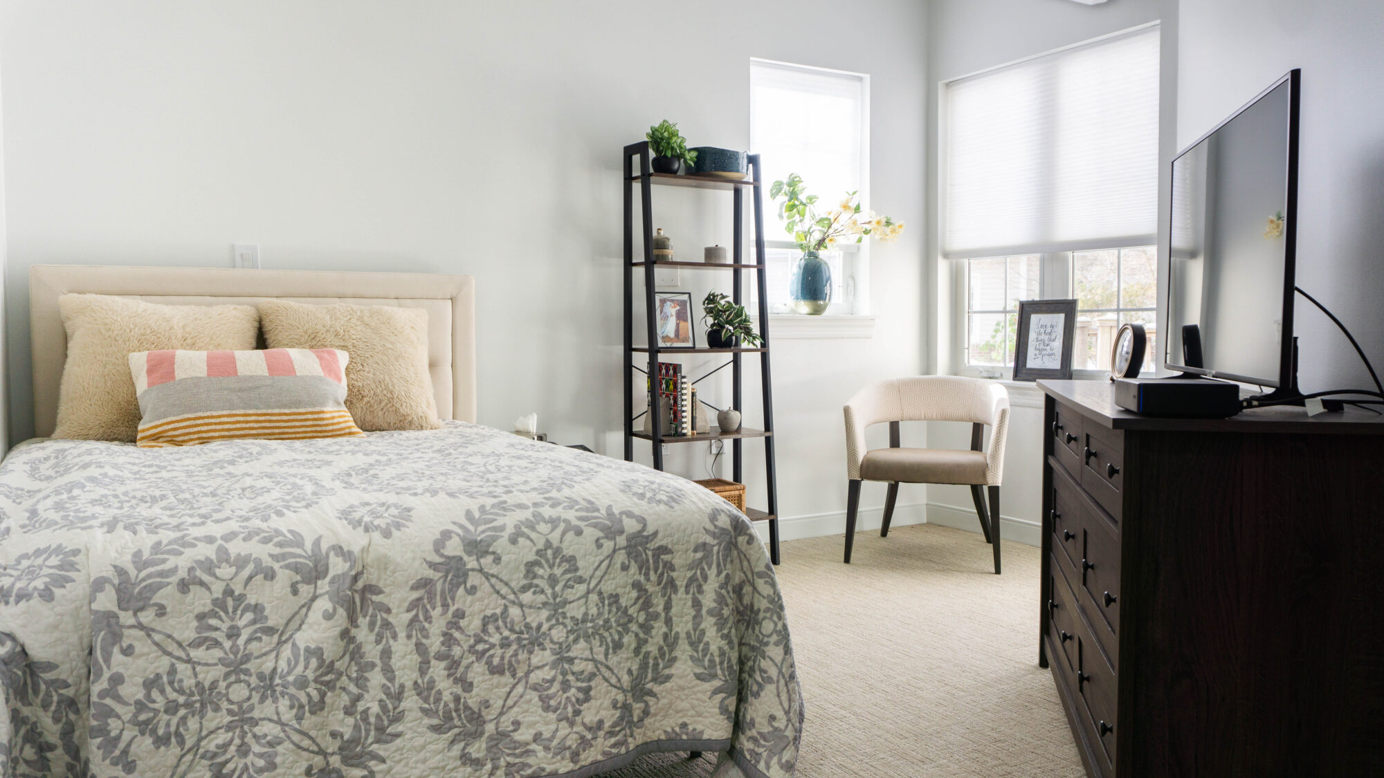 A warmly furnished bedroom with a neatly made bed featuring neutral-toned pillows and a patterned quilt. A modern shelving unit with plants, books, and personal touches stands by a window. A soft armchair and a dresser with a television complete the inviting, light-filled space.