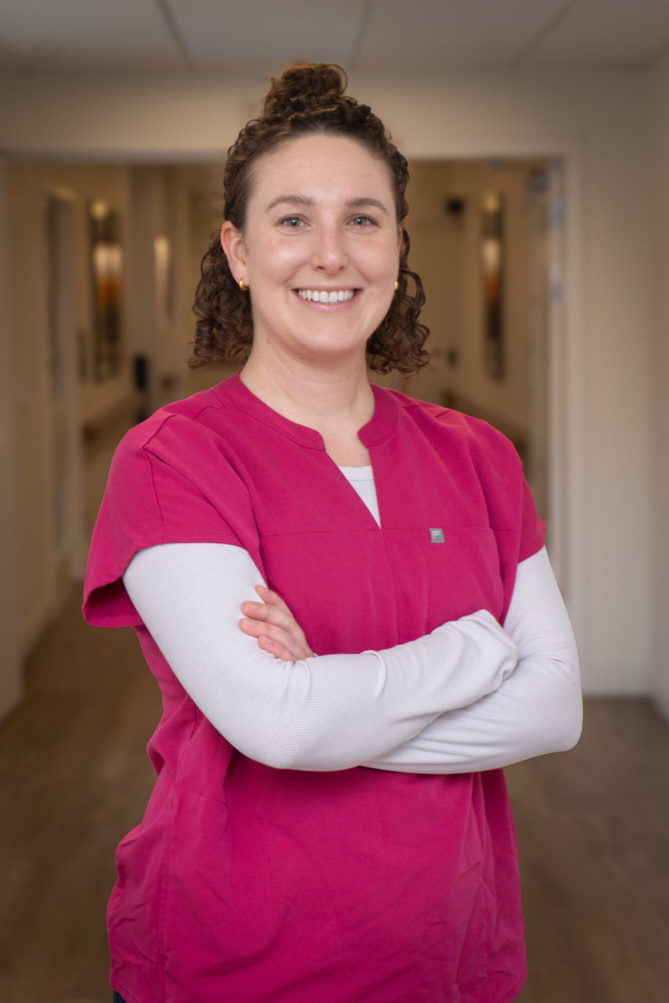 a white woman with brown hair wearing pink nursing scrubs stands in a white hallway with her arms crossed.